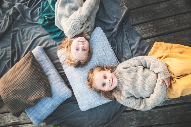 May include: Two children laying on a gray blanket with pillows. Both children are wearing gray sweaters and have long blonde hair. One child is wearing a green skirt and the other is wearing a yellow skirt.