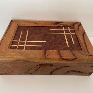 May include: A rectangular wooden box with a hinged lid. The box is made of two types of wood, a lighter wood for the frame and a darker wood for the top and sides. The top has a decorative inlay of thin, light-colored wood strips.