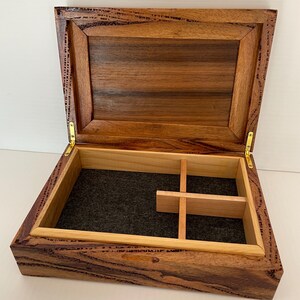 May include: Open wooden box with a hinged lid, featuring a dark brown exterior and a lighter brown interior. The box has a felt-lined base and wooden dividers. Gold-colored hinges secure the lid.