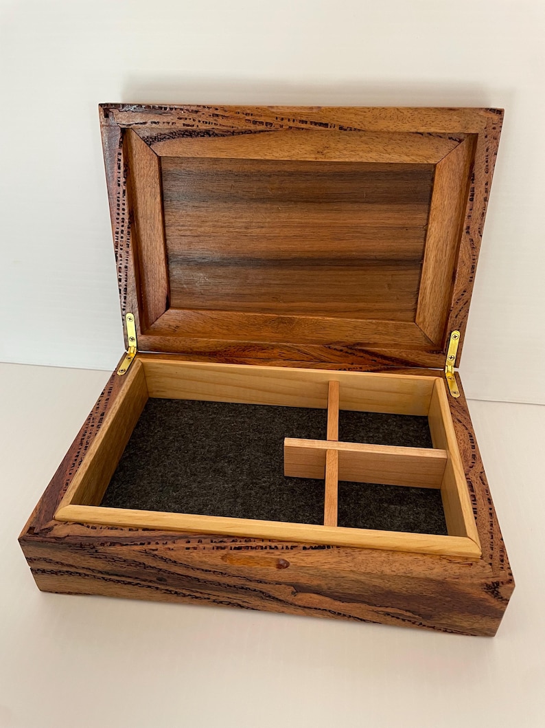 May include: Open wooden box with a hinged lid, featuring a dark brown exterior and a lighter brown interior. The box has a felt-lined base and wooden dividers. Gold-colored hinges secure the lid.
