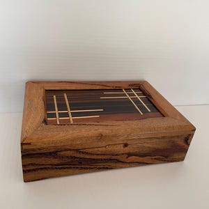 May include: A handcrafted wooden box with a rectangular shape. The lid features a geometric design with dark and light wood inlays. The box is made of a warm-toned wood with visible grain, showcasing a natural aesthetic.