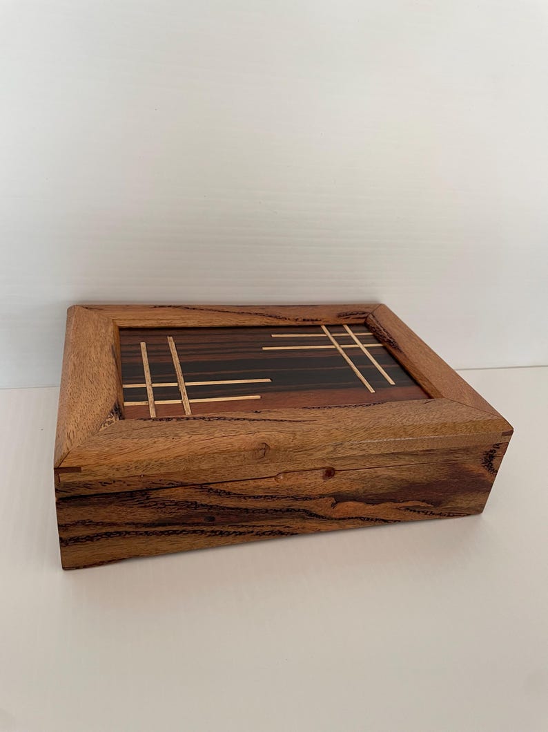 May include: A handcrafted wooden box with a rectangular shape. The lid features a geometric design with dark and light wood inlays. The box is made of a warm-toned wood with visible grain, showcasing a natural aesthetic.