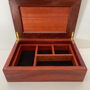 May include: Open wooden box with a hinged lid, crafted from reddish-brown wood. The interior features a black felt lining and several compartments. Gold-colored hinges secure the lid. The box is empty and sits on a white surface.