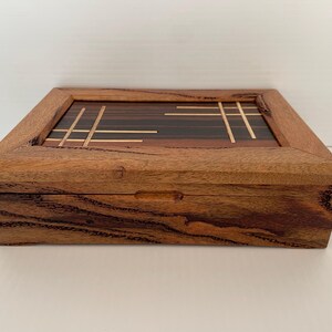 May include: A rectangular wooden box with a hinged lid. The box is made of a light brown wood with darker wood accents and a decorative inlaid top featuring a geometric design. The box has a simple, elegant design.