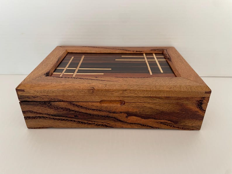 May include: A rectangular wooden box with a hinged lid. The box is made of a light brown wood with darker wood accents and a decorative inlaid top featuring a geometric design. The box has a simple, elegant design.