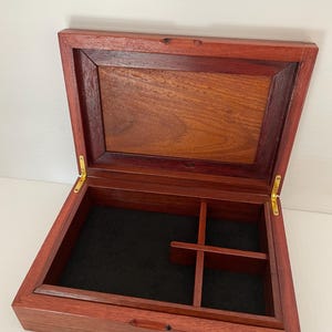 May include: Open wooden box with a hinged lid, crafted from reddish-brown wood. The interior is lined with black fabric and features dividers. The box is designed for storage and organization.