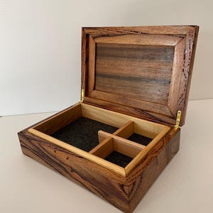 May include: Open wooden box with a hinged lid, featuring a dark brown exterior with lighter wood accents. The interior has a black felt lining and a divided compartment. The box is made of wood and has brass hinges.