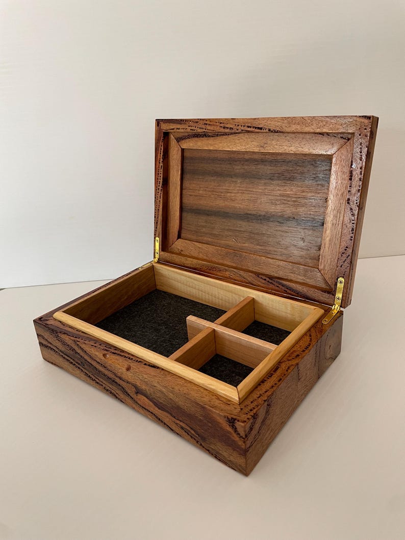 May include: Open wooden box with a hinged lid, featuring a dark brown exterior with lighter wood accents. The interior has a black felt lining and a divided compartment. The box is made of wood and has brass hinges.