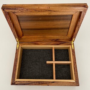 May include: Open wooden box with a hinged lid. The interior features a dark gray felt lining and a wooden divider creating compartments. The box is made of a reddish-brown wood with visible grain.