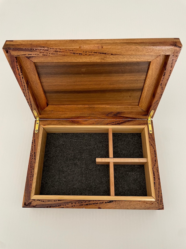May include: Open wooden box with a hinged lid. The interior features a dark gray felt lining and a wooden divider creating compartments. The box is made of a reddish-brown wood with visible grain.