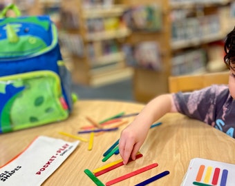 Peut inclure: Un enfant joue à un jeu avec des bâtonnets en bois colorés et une carte avec le numéro 10 dessus. La carte a une rangée de rectangles colorés dessus. L'enfant utilise les bâtonnets pour faire correspondre les couleurs sur la carte. Les bâtonnets sont disposés en un motif sur une table en bois. L'enfant porte un t-shirt gris avec un motif blanc. L'enfant est assis à une table dans une bibliothèque. Il y a un sac à dos bleu avec un dinosaure vert dessus en arrière-plan.