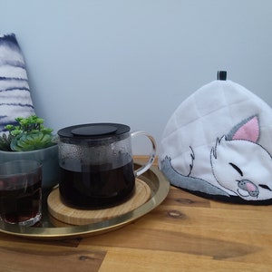 May include: A white teapot cosy with a grey and pink sleeping cat design. The cosy is on a wooden table next to a glass teapot filled with coffee and a glass of red liquid.