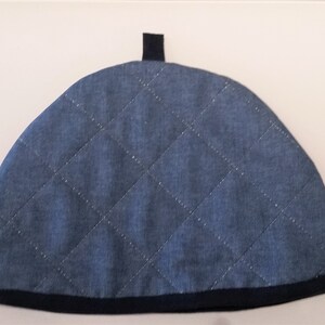 May include: A blue quilted tea cosy with a black trim and a black loop on top.