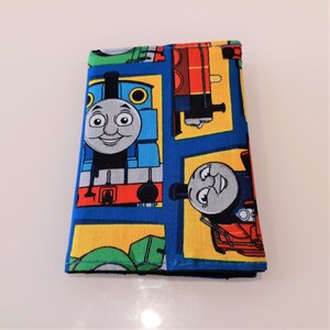 May include: A blue and yellow fabric wallet with a repeating pattern of Thomas the Tank Engine and his friends. The wallet features a variety of colours, including red, green, and blue.