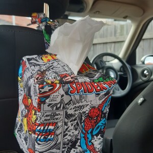 May include: A black and white comic book themed tissue box cover with a red and blue Spiderman graphic. The cover is attached to a car headrest with a black strap.