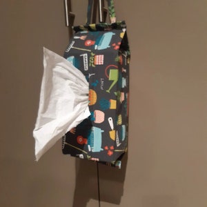 May include: A grey fabric tissue box holder with a colourful print of gardening tools, plants, and watering cans. The holder has a white tissue box inside and a black strap for hanging.