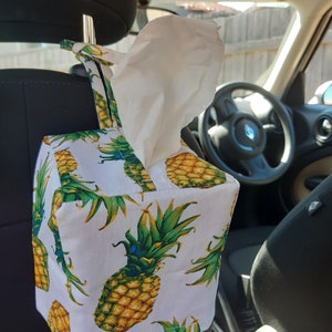 May include: A white tissue box cover with a pineapple pattern. The cover is hanging from the headrest of a car seat.