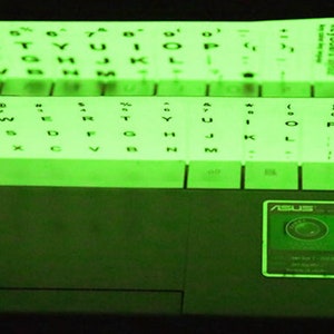 May include: A laptop keyboard with glowing green keyboard stickers applied. The stickers are arranged in rows and columns, with letters and symbols visible. The stickers are designed to illuminate in the dark. The laptop has an ASUS logo.