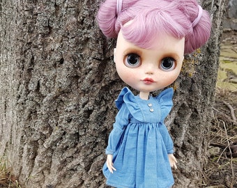 以下が含まれることがあります： ピンク色の髪を2つの団子に結い、フリル袖とボタンフロントの青いデニムドレスを着たブライスドール。人形は屋外に立っており、背景には木の幹があります。