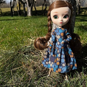 May include: A doll with long brown hair in braids wearing a blue and brown polka dot dress standing in a grassy area with trees in the background.