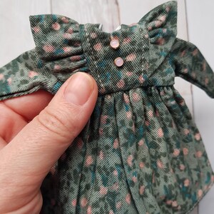 May include: A green and pink floral print dress with a ruffled collar and two gold buttons. The dress is made of a soft, lightweight fabric.