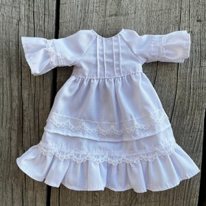 May include: A white doll dress with a tiered skirt and ruffled sleeves. The dress features delicate lace trim along the sleeves and skirt layers. The dress is made of a soft, lightweight fabric.