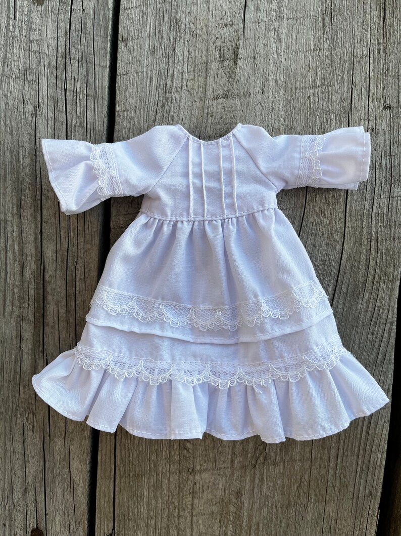 May include: A white doll dress with a tiered skirt and ruffled sleeves. The dress features delicate lace trim along the sleeves and skirt layers. The dress is made of a soft, lightweight fabric.