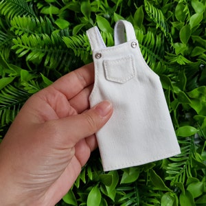 May include: A miniature white denim overall dress, featuring adjustable straps and a front pocket. The small garment has silver-coloured metal buttons and rivets. The overall is held against a backdrop of green foliage.