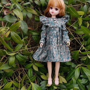 May include: A doll with long brown hair wearing a blue and pink floral dress with ruffled sleeves and pink shoes. The doll is standing in a bed of green leaves.
