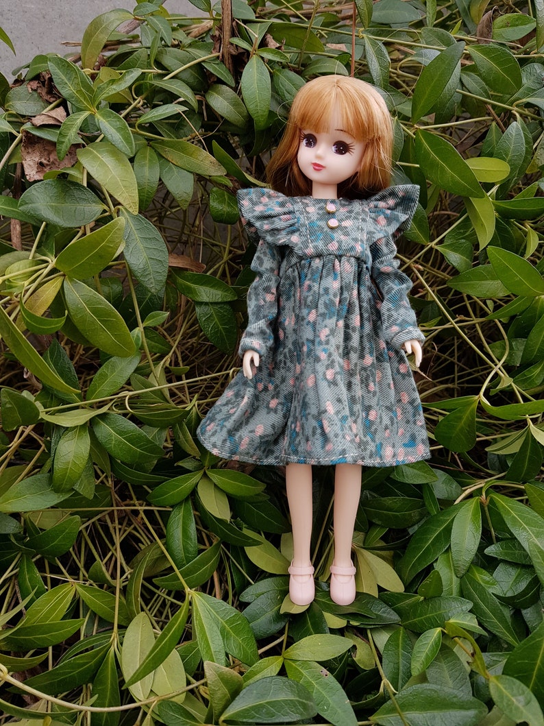 May include: A doll with long brown hair wearing a blue and pink floral dress with ruffled sleeves and pink shoes. The doll is standing in a bed of green leaves.