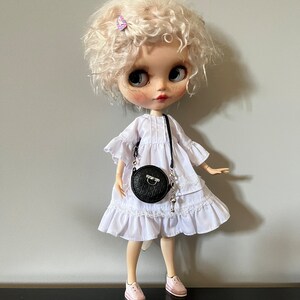 May include: A doll with blonde curly hair wearing a white dress with lace trim, pink sneakers, and a black round purse with a silver chain. A small purple butterfly hair clip is in the doll's hair. The doll has large blue eyes and freckles.