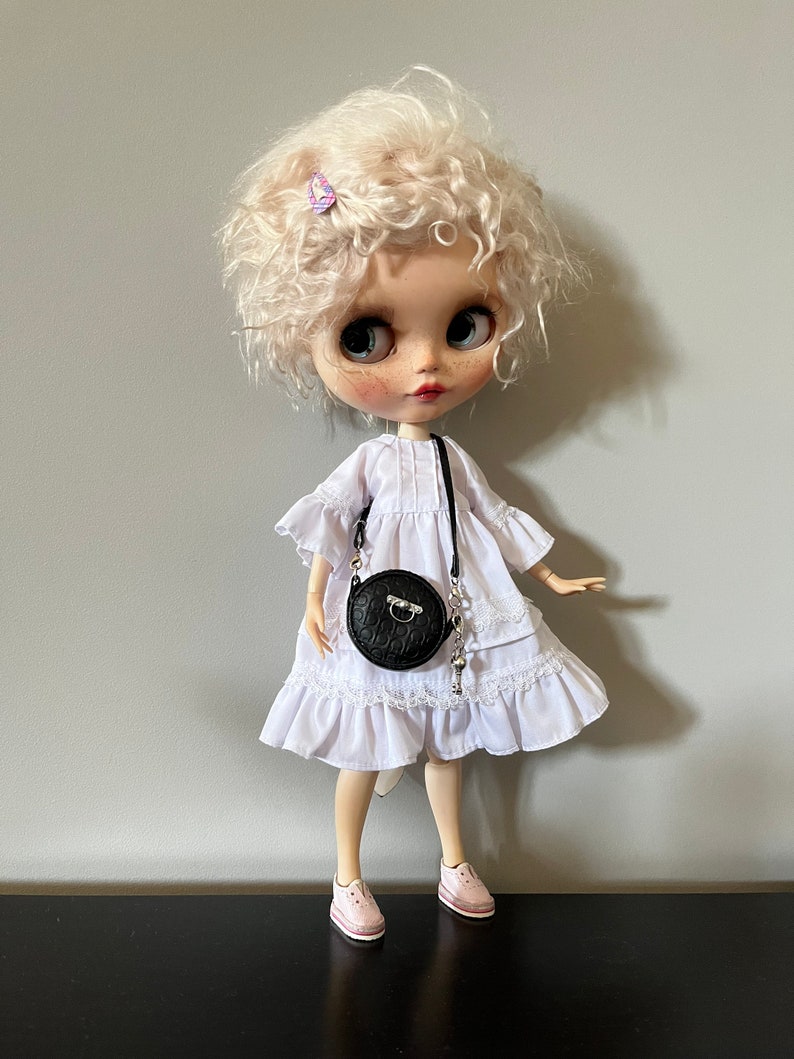 May include: A doll with blonde curly hair wearing a white dress with lace trim, pink sneakers, and a black round purse with a silver chain. A small purple butterfly hair clip is in the doll's hair. The doll has large blue eyes and freckles.