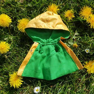 May include: A yellow and green hooded raincoat with a drawstring closure. The raincoat is lying on a bed of green grass with yellow dandelions.