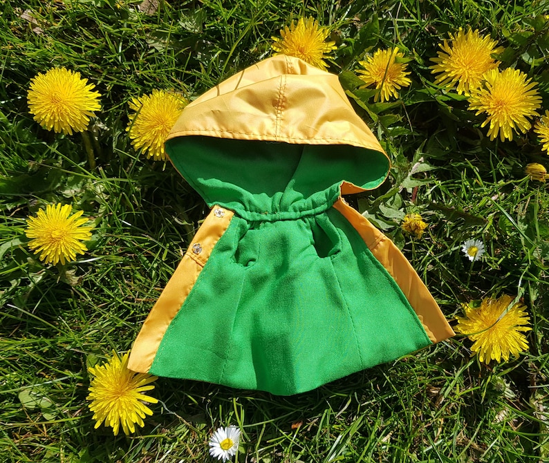 May include: A yellow and green hooded raincoat with a drawstring closure. The raincoat is lying on a bed of green grass with yellow dandelions.