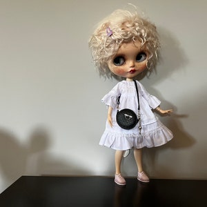 May include: A doll with curly blonde hair, wearing a white dress, pink shoes, and a black round purse. A small butterfly hair clip is in the doll's hair. The doll is standing on a black surface.