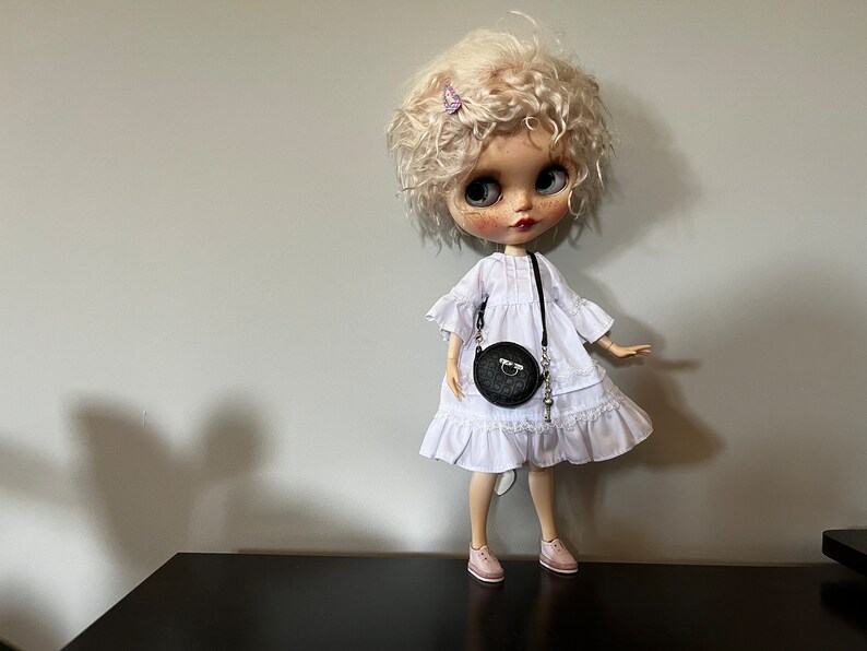 May include: A doll with curly blonde hair, wearing a white dress, pink shoes, and a black round purse. A small butterfly hair clip is in the doll's hair. The doll is standing on a black surface.