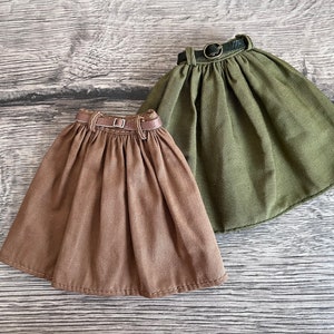 Könnte beinhalten: Zwei puppenkleine Röcke. Einer ist mittelbraun, der andere olivgrün. Beide Röcke haben ein plissiertes Design und einen braunen Gürtel mit Metallschnalle. Die Röcke sind aus weichem Stoff.