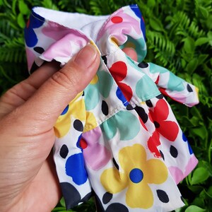 May include: A white fabric doll dress with a colorful floral print. The dress has a ruffled hem and a white collar.
