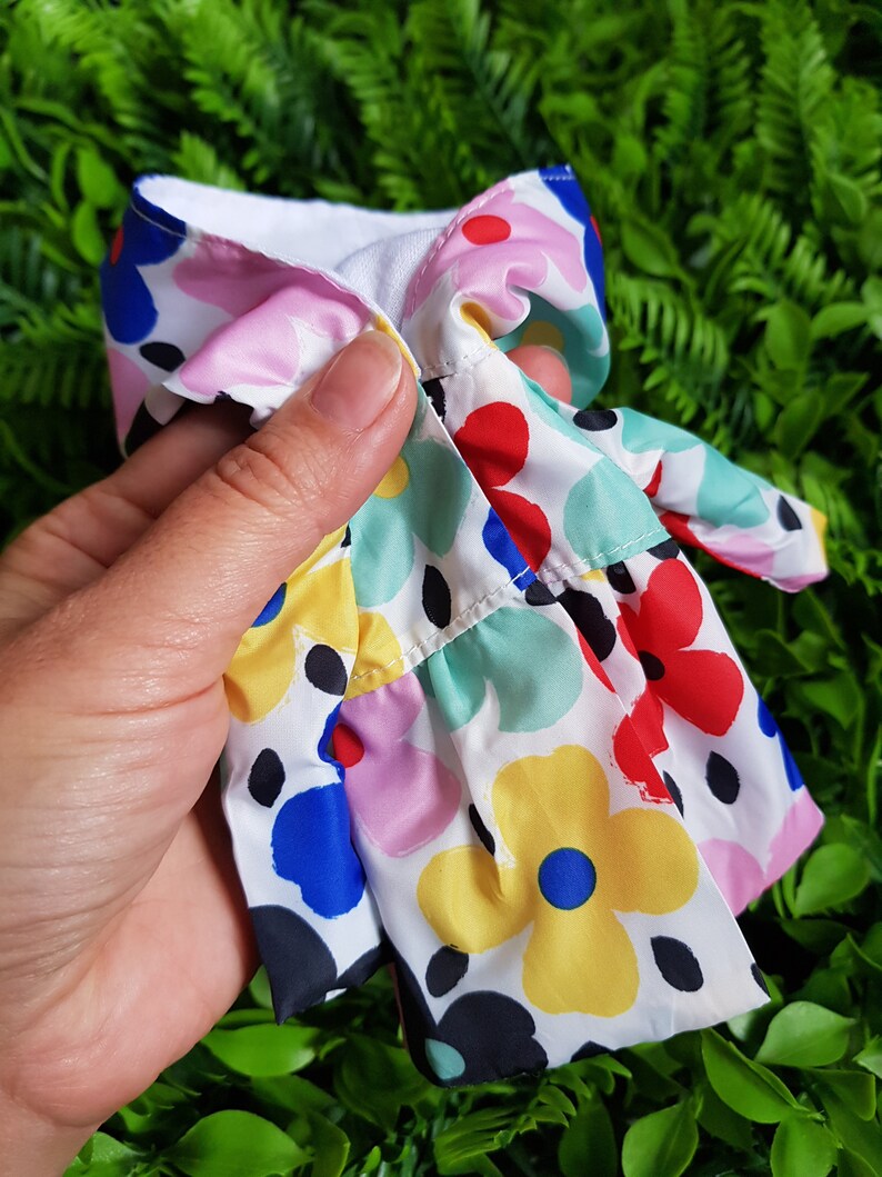 May include: A white fabric doll dress with a colorful floral print. The dress has a ruffled hem and a white collar.