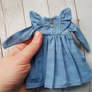May include: A light blue denim dress with long sleeves and a ruffled collar. The dress has two silver buttons at the neckline.