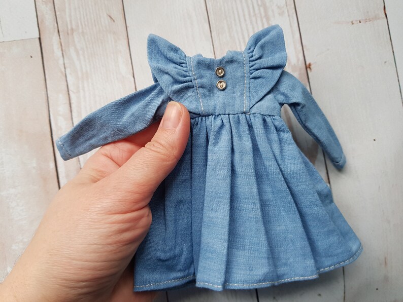 May include: A light blue denim dress with long sleeves and a ruffled collar. The dress has two silver buttons at the neckline.