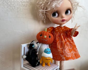 May include: A Blythe doll in an orange dress with white polka dots and pumpkin patterns. Beside the doll is a small white chair with a black cat, a pumpkin, and a yellow and blue figure.