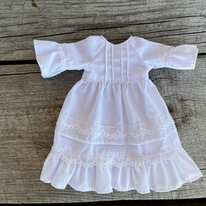 May include: A white doll dress with a round neckline, short sleeves, and a tiered skirt. The dress features delicate lace trim on the sleeves and skirt layers. The fabric appears to be lightweight and suitable for a doll.