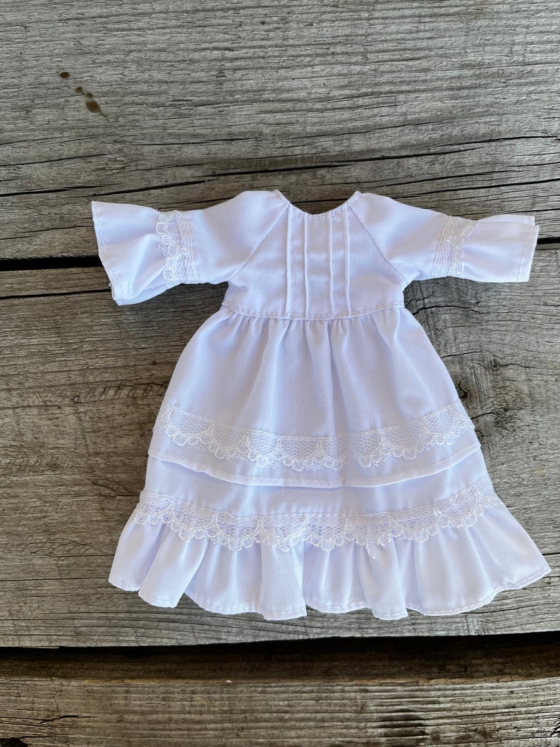 May include: A white doll dress with a round neckline, short sleeves, and a tiered skirt. The dress features delicate lace trim on the sleeves and skirt layers. The fabric appears to be lightweight and suitable for a doll.