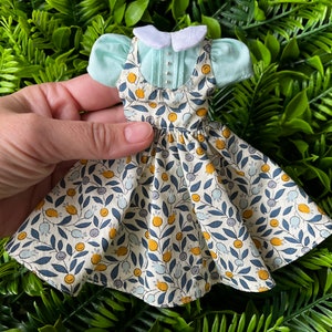 May include: A doll-sized dress with a white collar and a floral print featuring yellow and blue flowers on a white background. The dress is paired with a light blue blouse with a white collar.