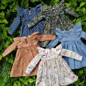 May include: Five doll dresses in various floral patterns and colors, including blue, green, brown, and white. The dresses have ruffled collars and long sleeves.
