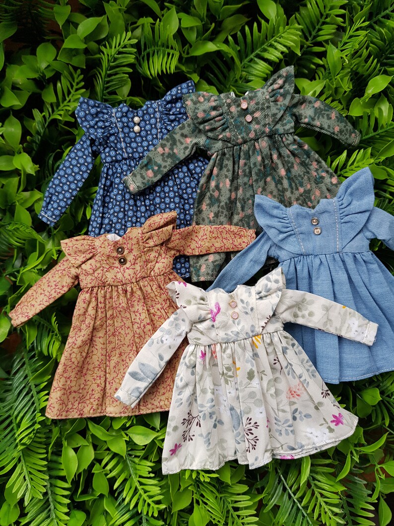 May include: Five doll dresses in various floral patterns and colors, including blue, green, brown, and white. The dresses have ruffled collars and long sleeves.