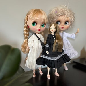 May include: Three fashion dolls are posed on a dark surface. The dolls have large eyes and wear dresses in white and black. The doll in the center wears a black dress with white lace trim. The doll on the left has blonde braids and a white dress. The doll on the right has curly blonde hair and a white dress.