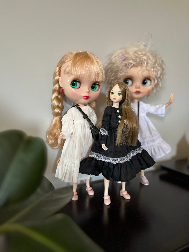 May include: Three fashion dolls are posed on a dark surface. The dolls have large eyes and wear dresses in white and black. The doll in the center wears a black dress with white lace trim. The doll on the left has blonde braids and a white dress. The doll on the right has curly blonde hair and a white dress.