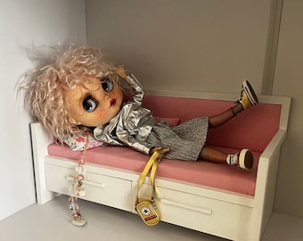Puede incluir: Una muñeca con el pelo rubio rizado está tumbada en un sofá en miniatura rosa y blanco. La muñeca lleva una chaqueta plateada, una falda gris y medias a rayas blancas y negras. La muñeca lleva zapatillas negras y doradas. El sofá tiene dos cajones.